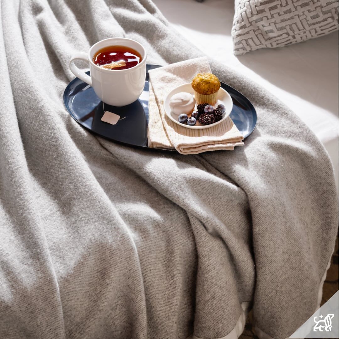 Cozy scene with a steaming mug of tea, fresh muffin, berries, and marshmallow on a blue tray atop a soft, gray blanket-covered bed.