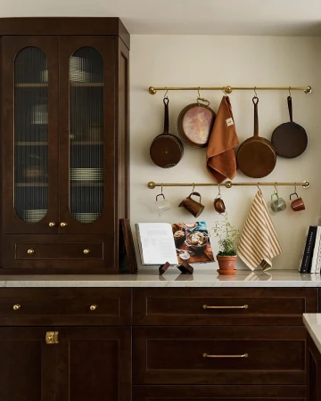 Warm, inviting kitchen scene with dark wood cabinetry, brass hardware, hanging copper cookware, and a cookbook displayed on the counter.
