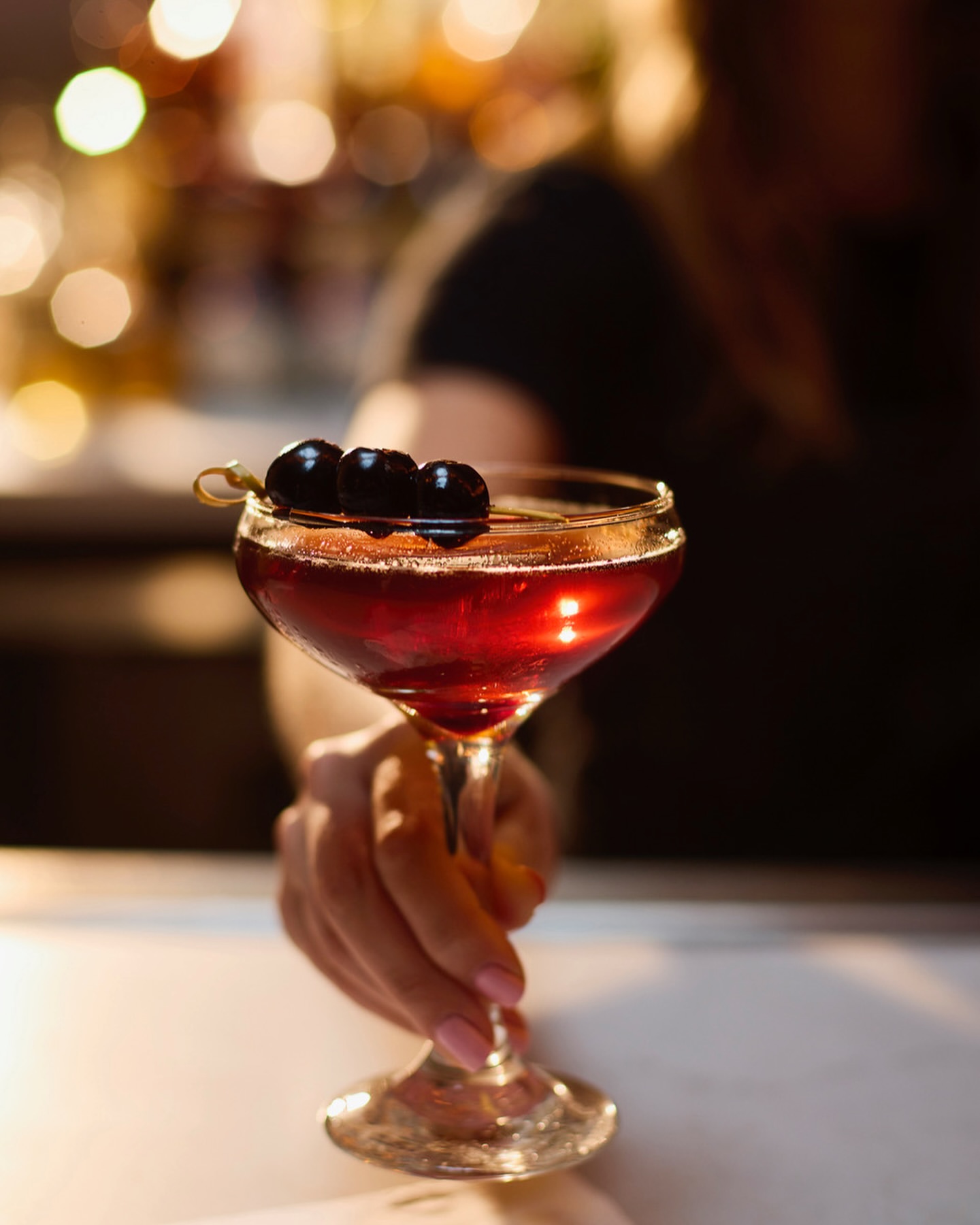 Cocktail in coupe glass held by a woman's manicured hand with three dark cherries, against a bokeh-lit bar background.