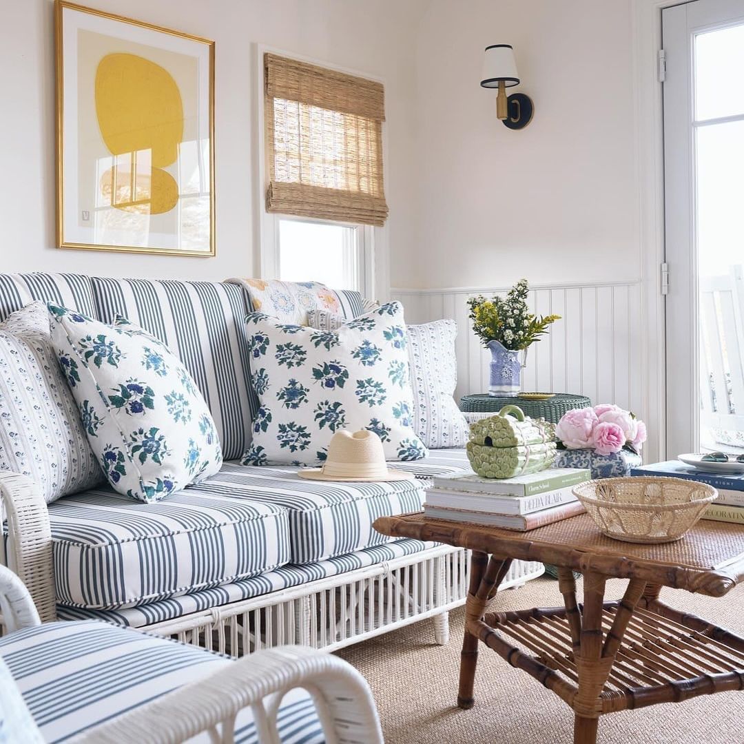 Living room interior with white wicker sofa and armchairs, floral pillows, rattan coffee table, framed art, and natural light from windows.
