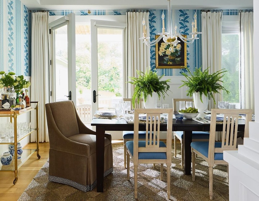 Formal dining room interior with a dark wood table, upholstered chairs, large ferns, blue patterned wallpaper, and natural light from open doors.