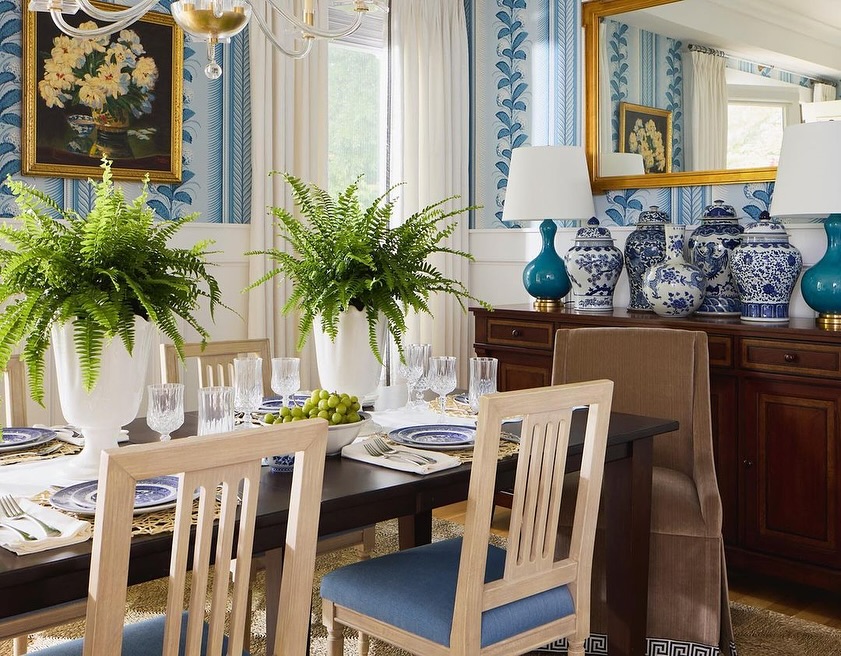 Dining room interior featuring a table set with dishes and glassware, flanked by ferns, beneath a chandelier and framed painting.