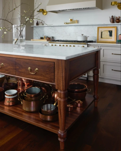 Kitchen interior with a wood island, marble countertop, copper pots, and white cabinets, creating a warm, traditional culinary space.