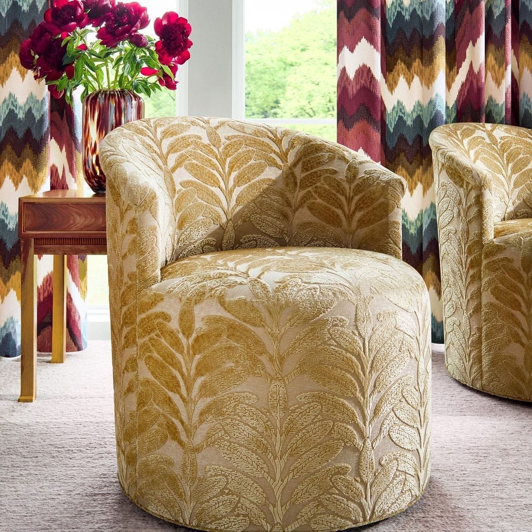 Interior with patterned armchairs, a wood side table, and a glass vase with red flowers, set against a backdrop of patterned curtains.