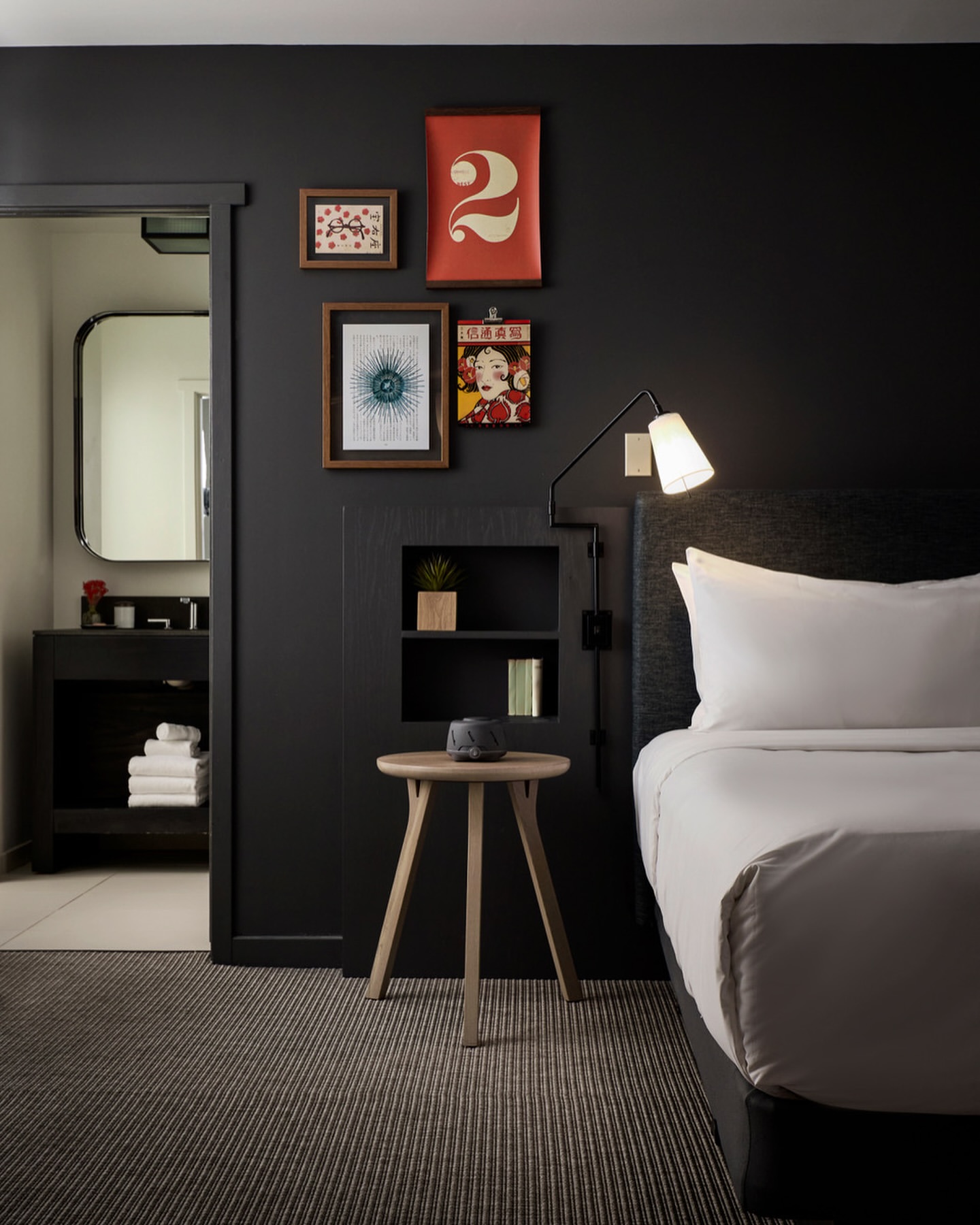 Modern hotel room interior with a bed, small side table, and a doorway to a bathroom, featuring a dark colour scheme and minimalist decor.