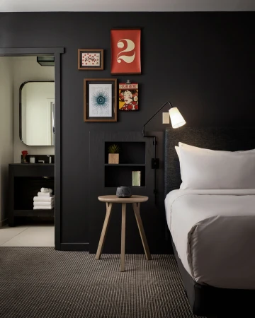 Modern hotel room interior with a bed, small side table, and a doorway to a bathroom, featuring a dark colour scheme and minimalist decor.