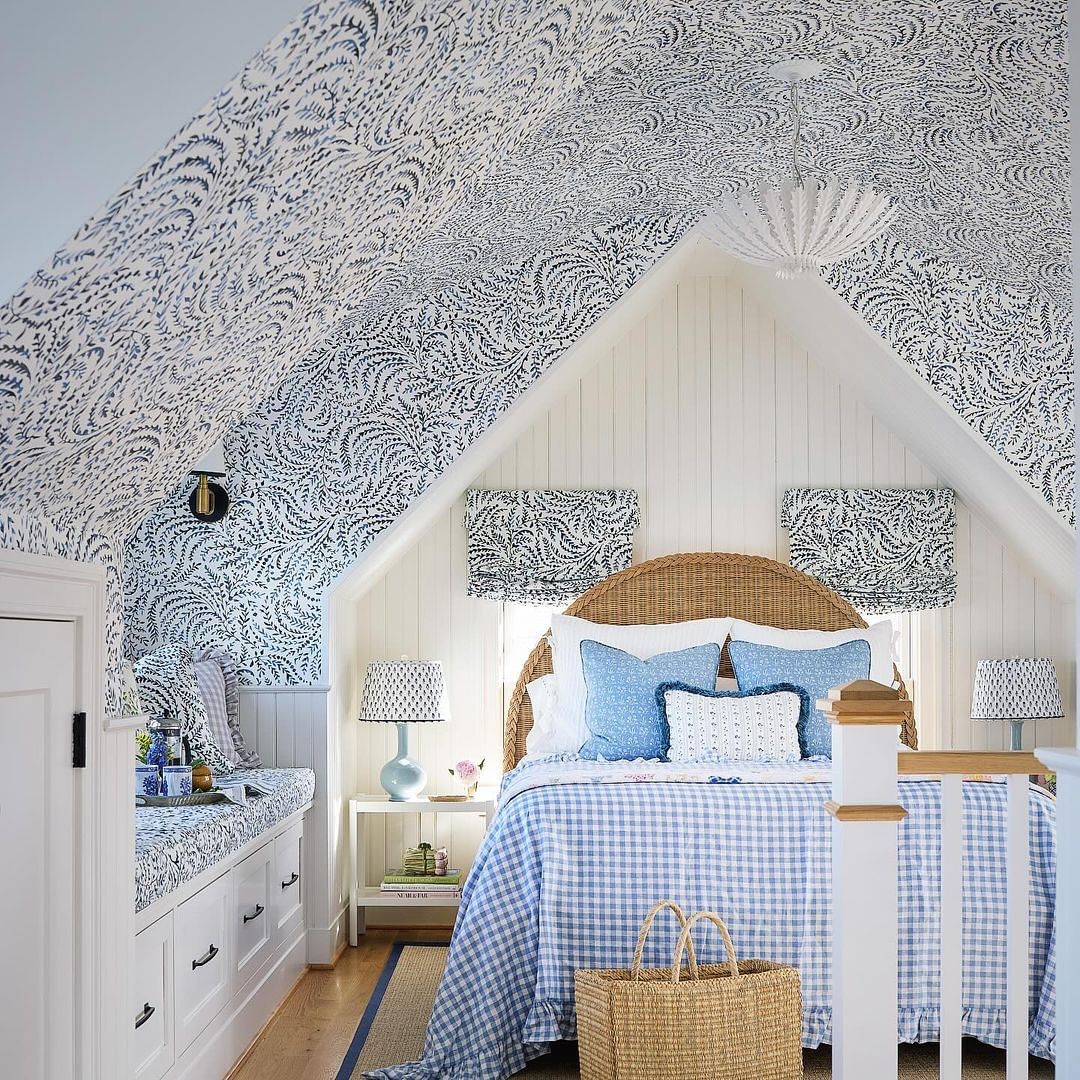 Attic bedroom interior with floral wallpaper, white wood paneling, wicker headboard, a bed with a blue gingham cover, and natural light.