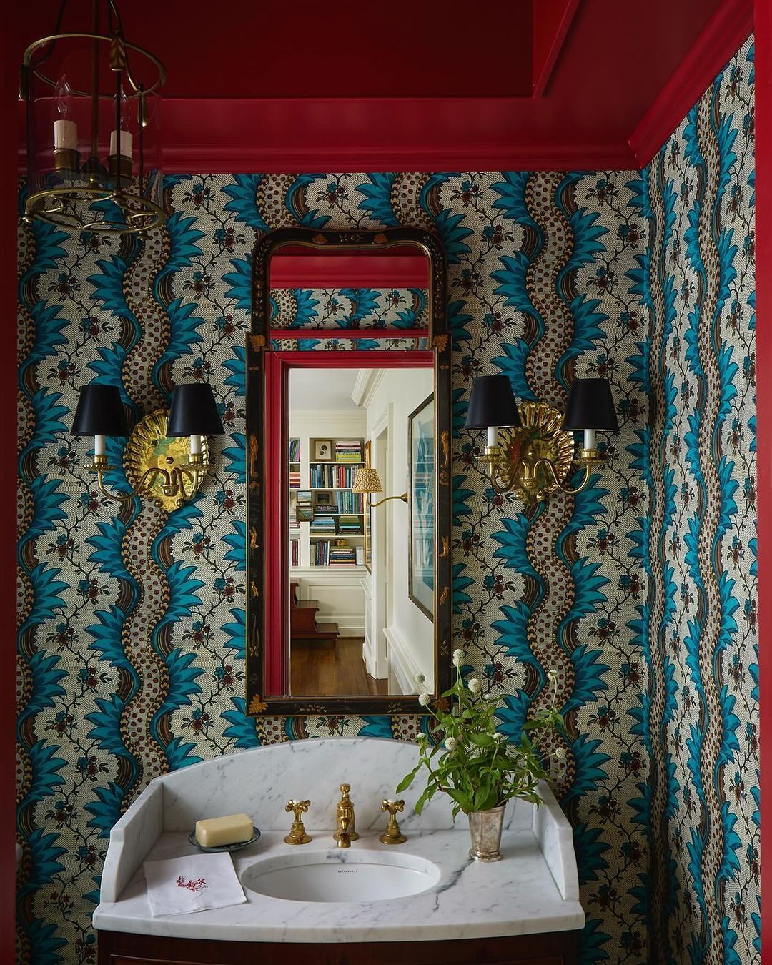 Bathroom interior featuring a sink, ornate wallpaper, sconces, mirror, and red trim, creating a visually rich and traditionally styled space.