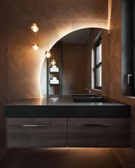 Contemporary bathroom features textured wood wall, curved mirror with lights, floating vanity with gray countertop and dark wood drawers.
