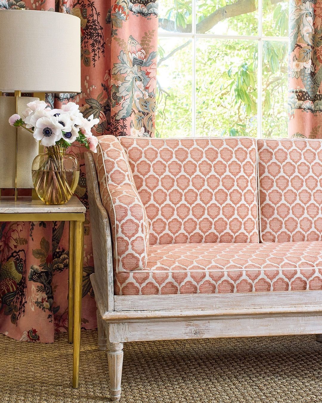 Living room interior with a coral patterned sofa near a side table, flowers in a vase, and patterned coral curtains in front of a window.
