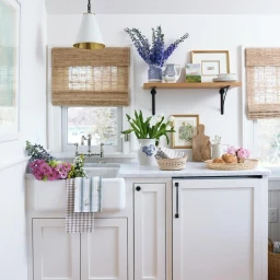 Bright kitchen interior featuring white cabinetry, a farmhouse sink with fresh flowers, and woven bamboo window blinds by Kip Dawkins.