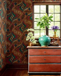 Potted plants on a rustic reddish-brown wooden chest of drawers against colorful patterned wallpaper and a multi-pane window.