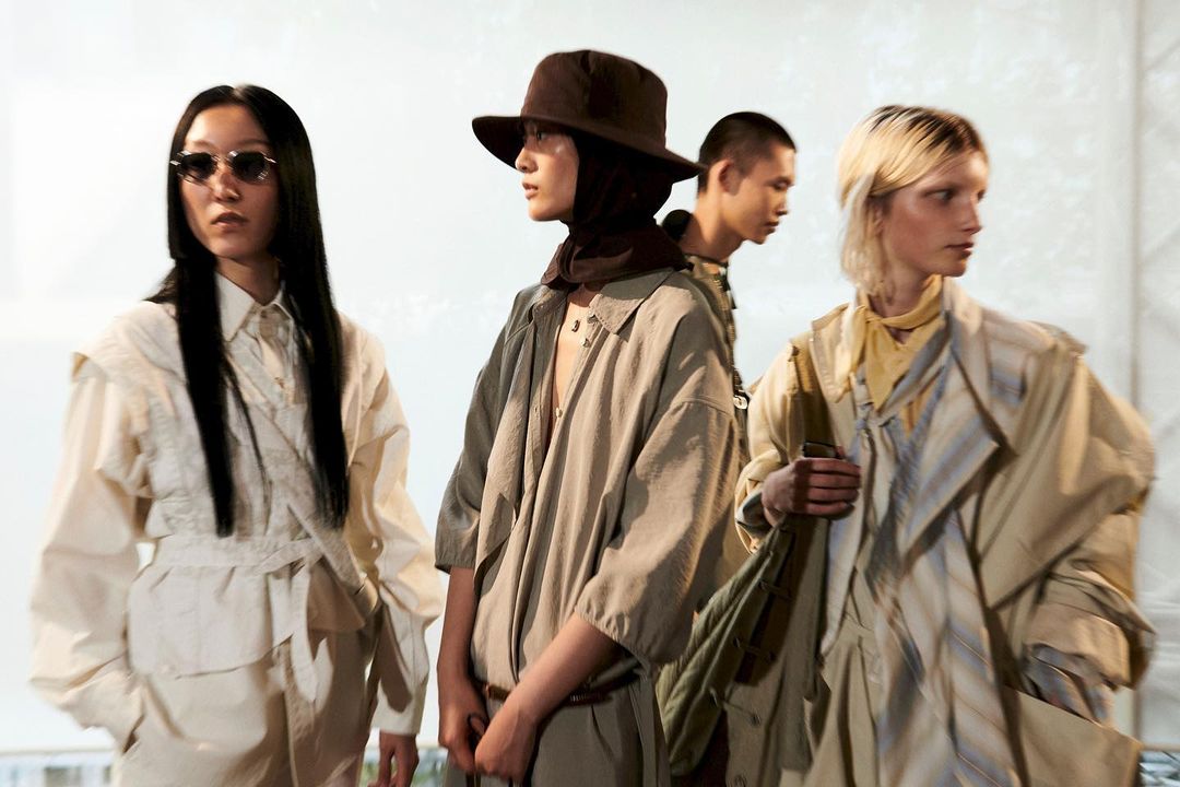 Four fashion models in neutral-toned outfits pose indoors under bright lighting, showcasing earthy apparel and diverse styles with elegance.