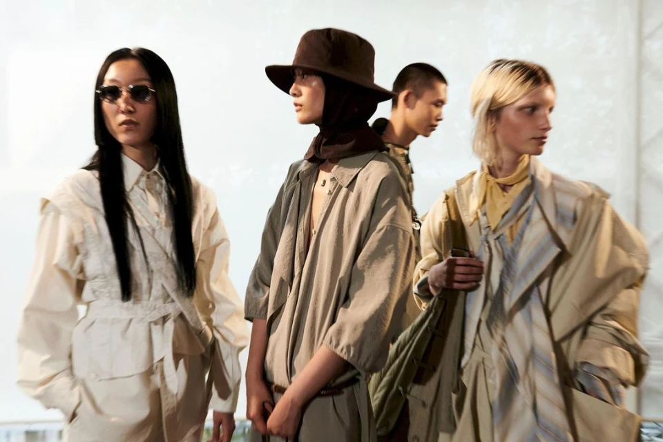 Four fashion models in neutral-toned outfits pose indoors under bright lighting, showcasing earthy apparel and diverse styles with elegance.