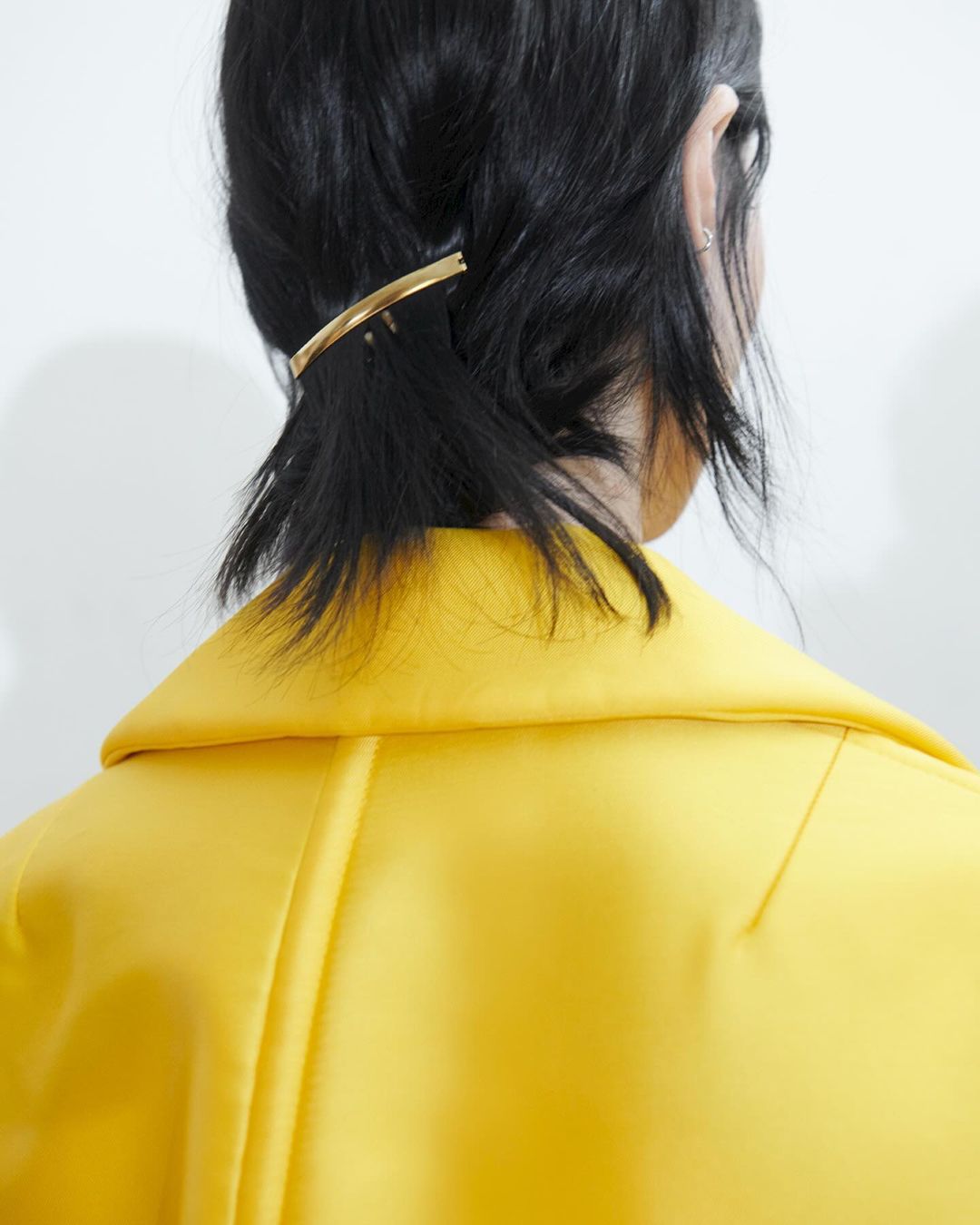 A woman from behind with short black hair pinned with a gold barrette, wearing a bright yellow jacket against a white background.