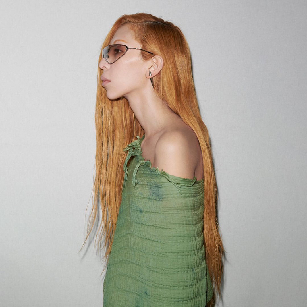 Profile shot of a slender adult female with long red-orange hair, wearing sunglasses and a green distressed fabric wrap, against a white wall.