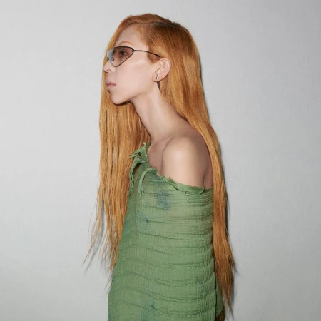 Profile shot of a slender adult female with long red-orange hair, wearing sunglasses and a green distressed fabric wrap, against a white wall.