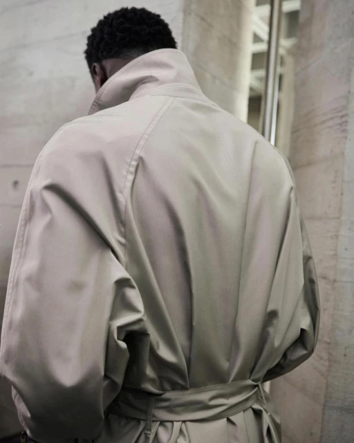 Back view of a Black man wearing a beige trench coat with a tied belt, standing indoors near a textured beige wall and metal structure.