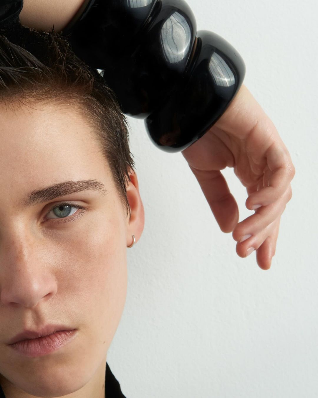 Close-up portrait of a woman with short hair wearing a large black sculptural bangle bracelet against a plain white backdrop.