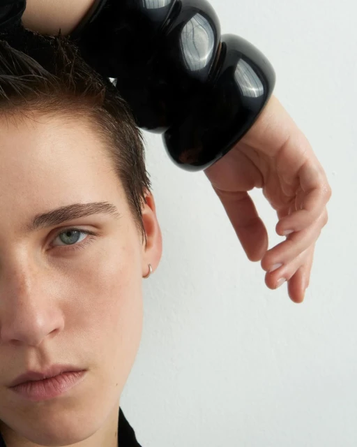 Close-up portrait of a woman with short hair wearing a large black sculptural bangle bracelet against a plain white backdrop.