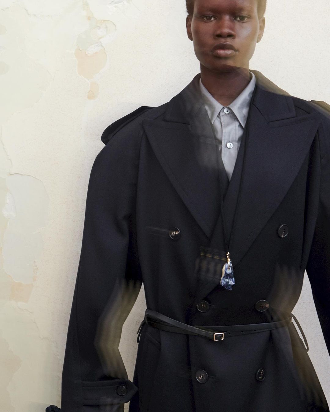 Androgynous model poses indoors wearing black trench coat, dress shirt, and pendant against a textured neutral wall in a fashion editorial.