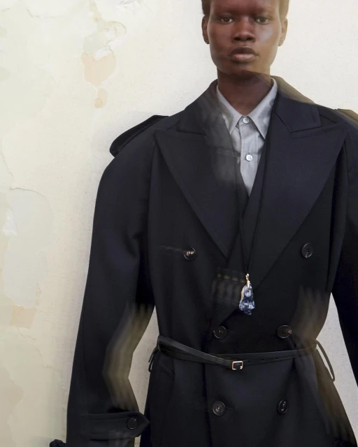 Androgynous model poses indoors wearing black trench coat, dress shirt, and pendant against a textured neutral wall in a fashion editorial.
