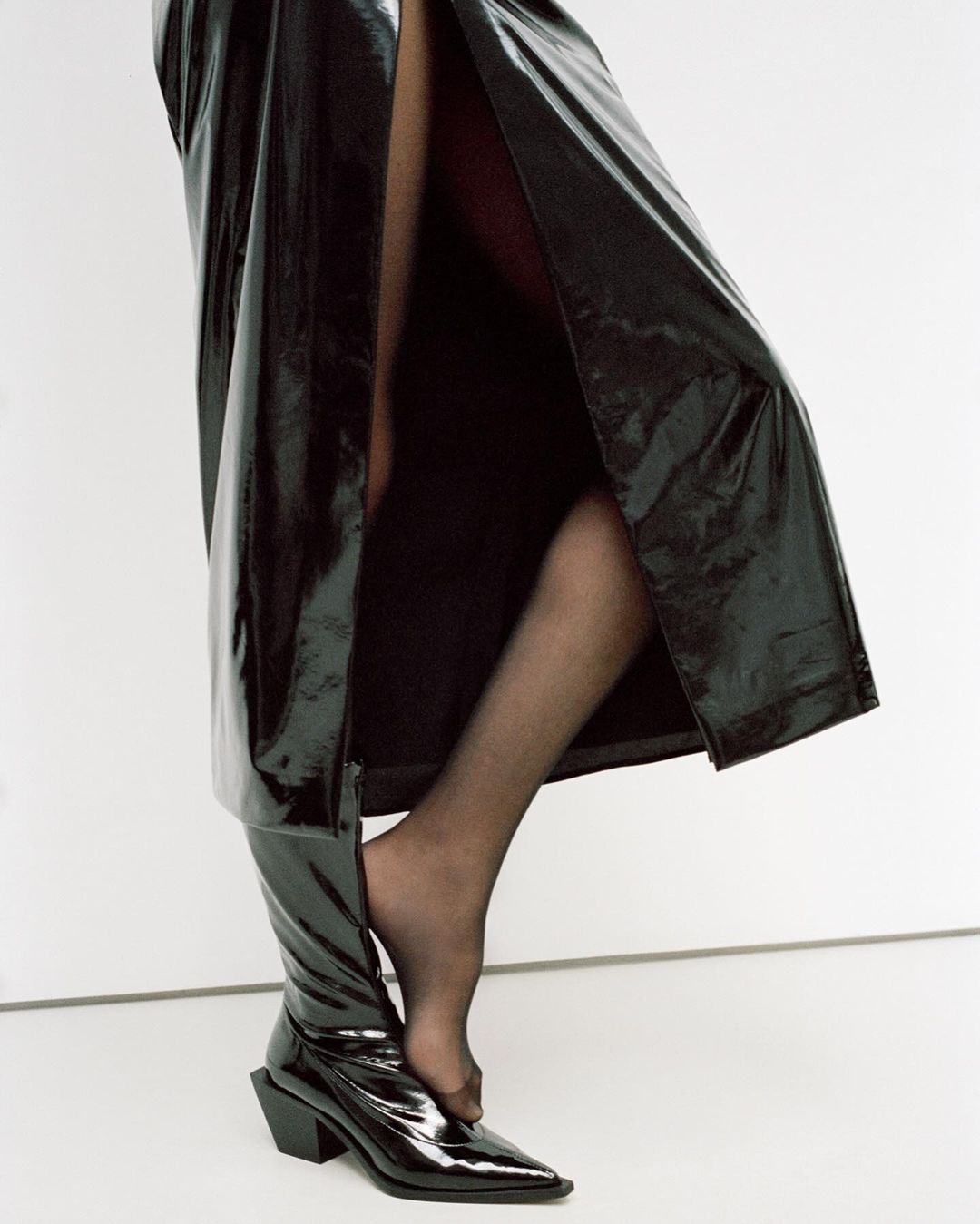 Partial view of adult female wearing black glossy over-the-knee boot and long black shiny coat in a vertical studio fashion shot.