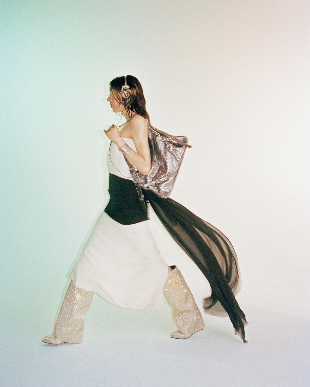 Fashionable adult woman in long white dress, sequined boots, silver shoulder bag, and headphones poses in a brightly lit studio.