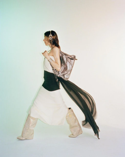 Fashionable adult woman in long white dress, sequined boots, silver shoulder bag, and headphones poses in a brightly lit studio.