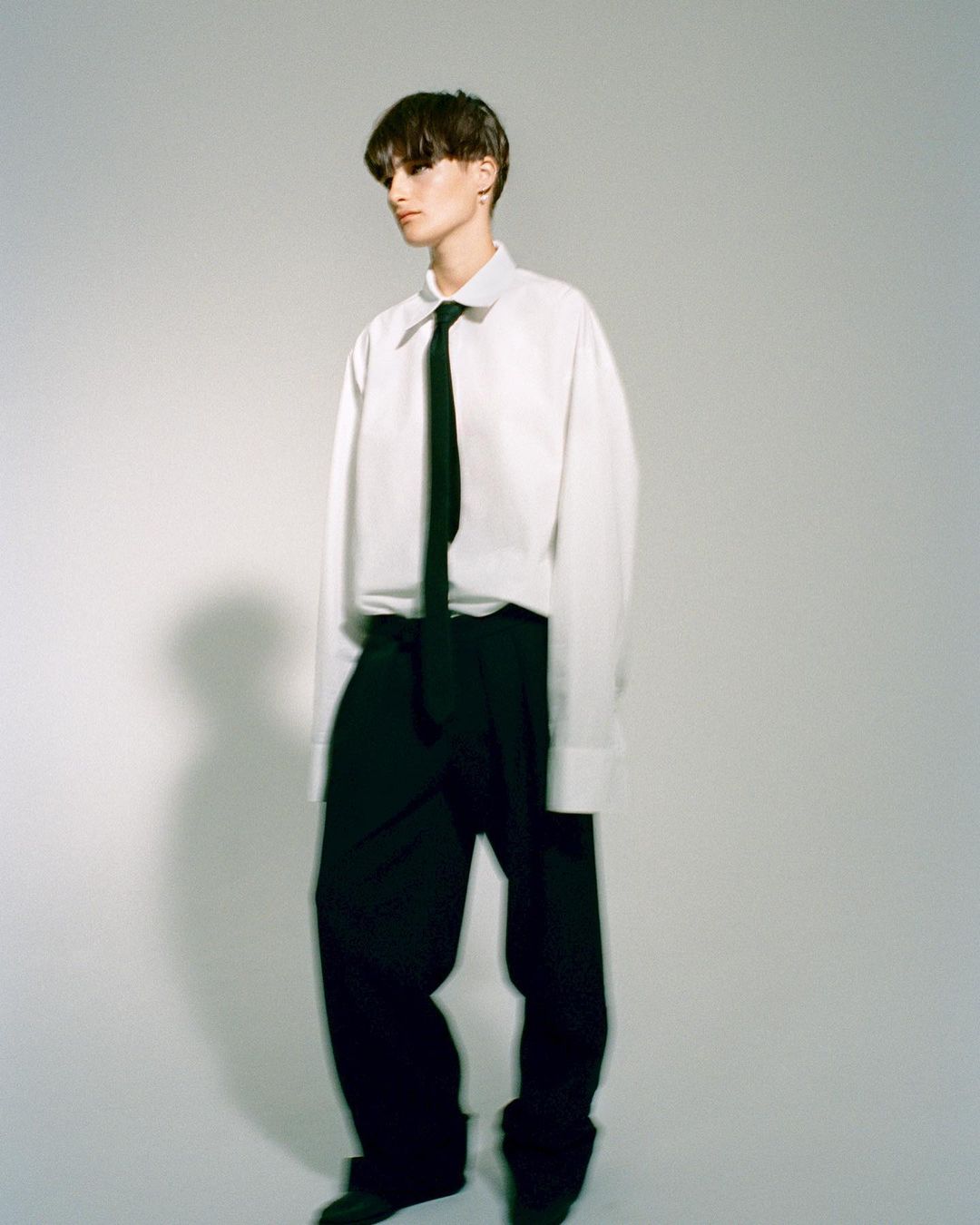 Stylish person with short dark hair poses in a white button-down shirt, black tie, and trousers against a neutral backdrop.
