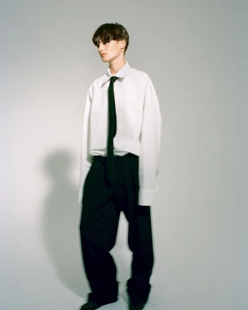 Stylish person with short dark hair poses in a white button-down shirt, black tie, and trousers against a neutral backdrop.