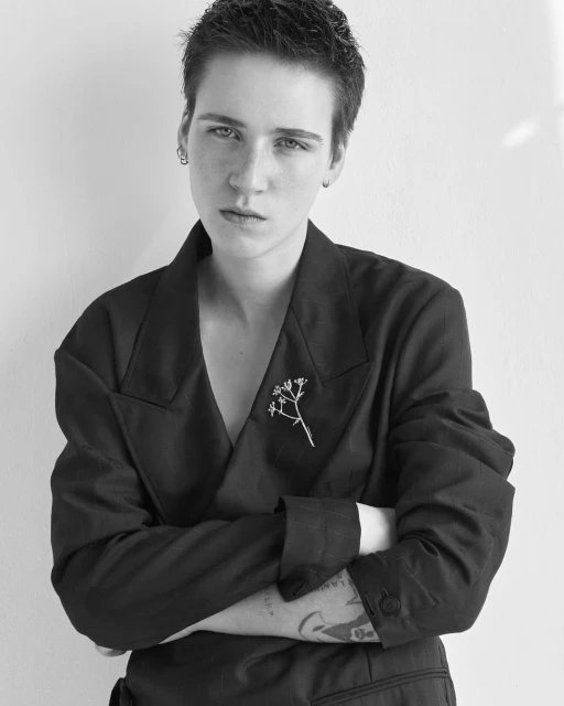 Monochrome portrait of a young adult with short hair in a dark jacket, arms crossed, and a neutral expression against a white wall.