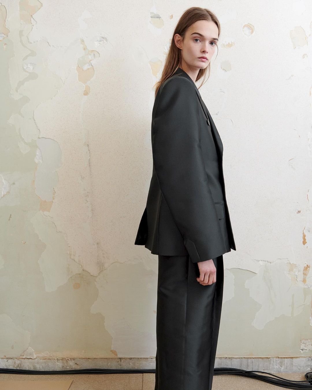 Woman in an olive-green, oversized suit stands in a studio against a cracked, cream-colored wall with visible wiring at the bottom.