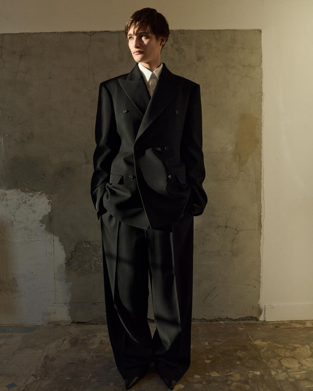 Adult male poses indoors wearing an oversized double-breasted black suit with peaked lapels against a mottled grey backdrop.