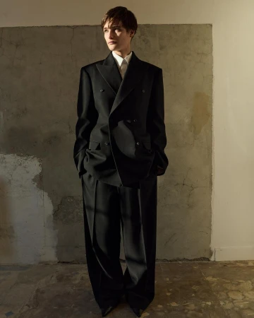 Adult male poses indoors wearing an oversized double-breasted black suit with peaked lapels against a mottled grey backdrop.