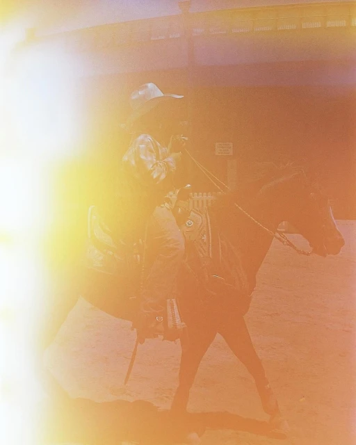 Man riding a horse indoors, wearing a cowboy hat and a patterned shirt, with bright lighting casting a warm glow.