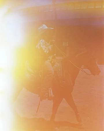 Man riding a horse indoors, wearing a cowboy hat and a patterned shirt, with bright lighting casting a warm glow.