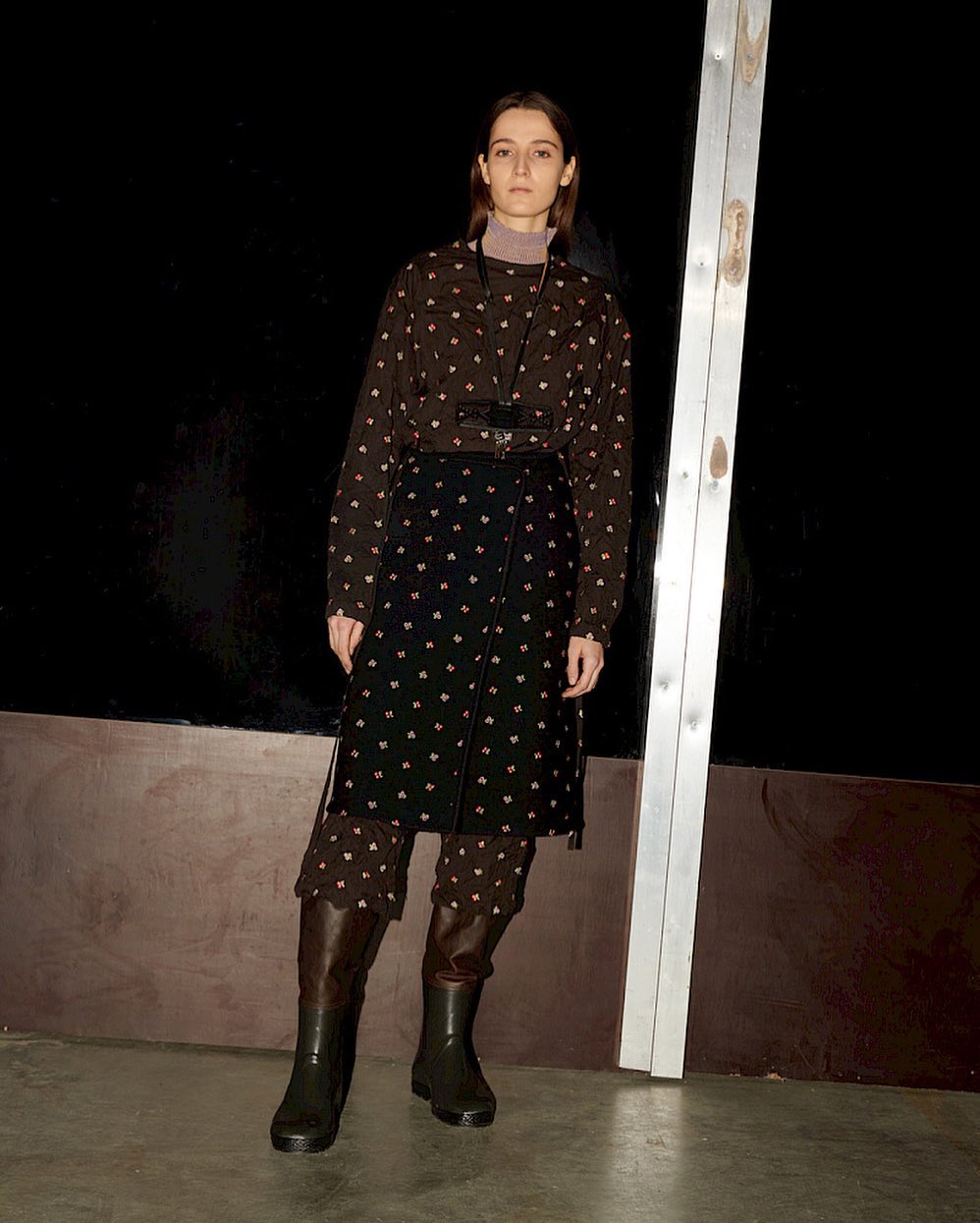 Fashionable young woman poses in a patterned shirt, skirt, and brown boots against a dark background and industrial metal support.