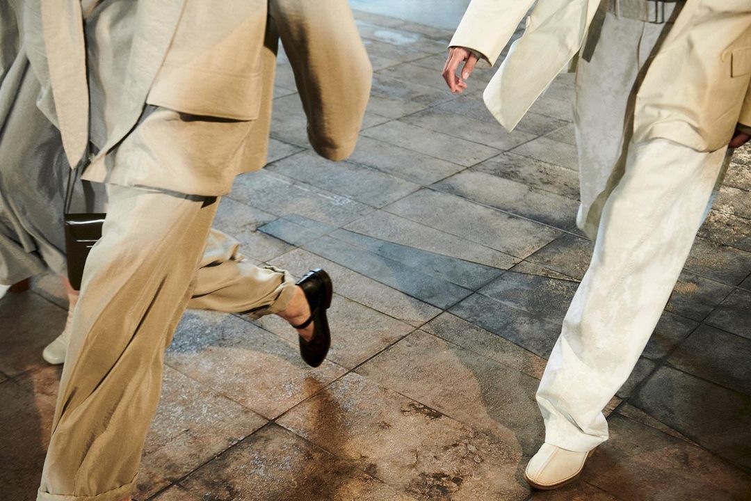 Close-up fashion shot of two people walking on a tiled floor wearing beige and white pants, shoes, and jackets, neutral colour palette.