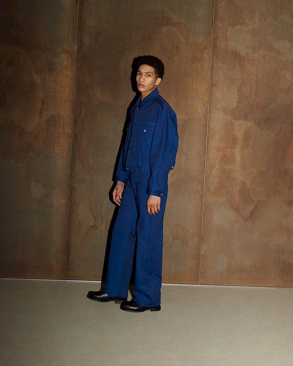 A young adult poses in a dark blue denim suit against a textured brownish wall in a fashion style shot. Light comes from the side.
