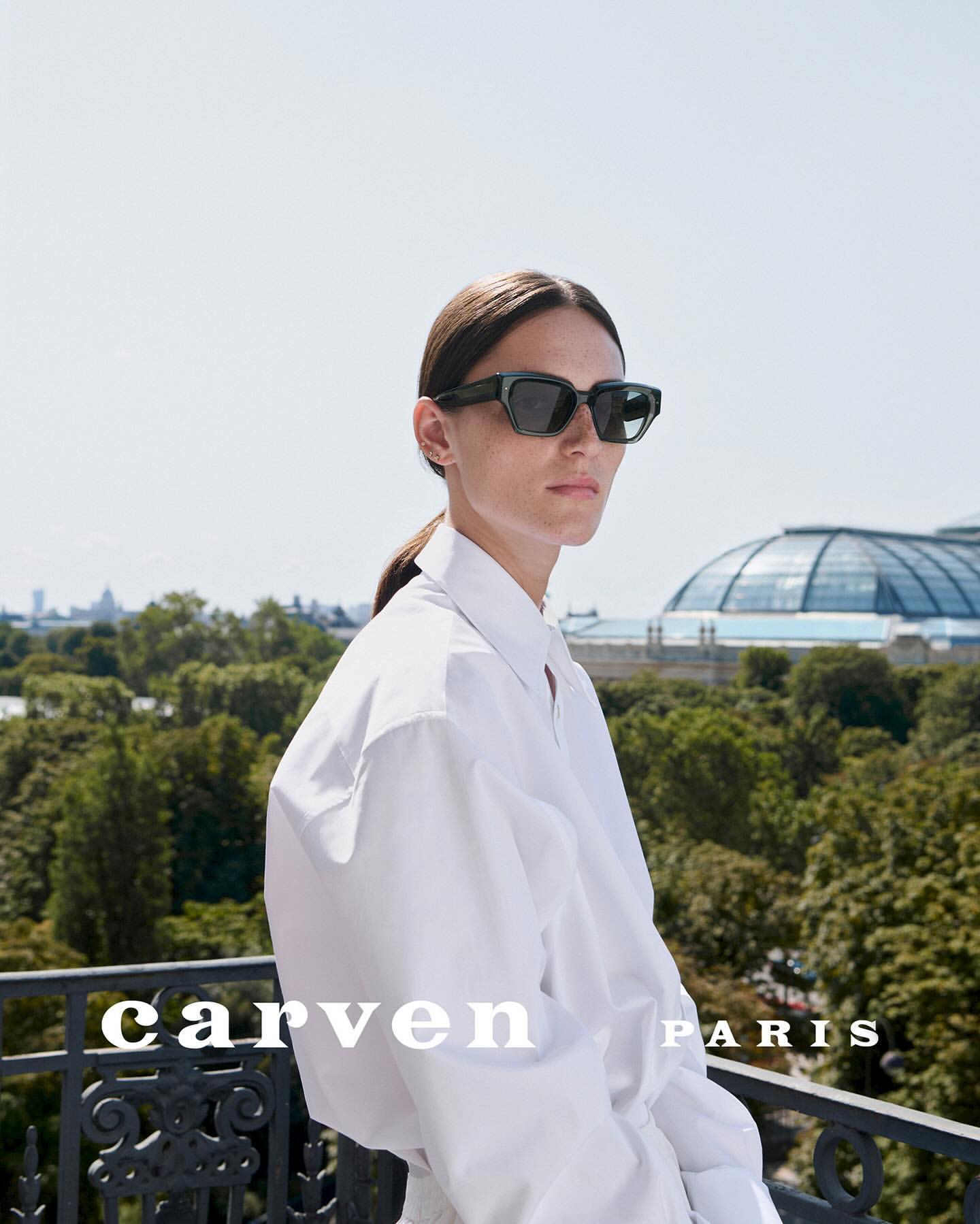 A woman wears dark sunglasses and a white shirt outdoors, posing on a balcony with trees and a large glass-domed building visible.