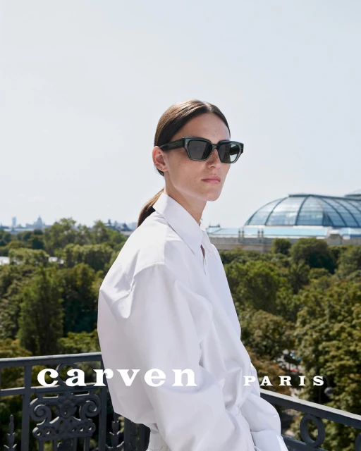 A woman wears dark sunglasses and a white shirt outdoors, posing on a balcony with trees and a large glass-domed building visible.