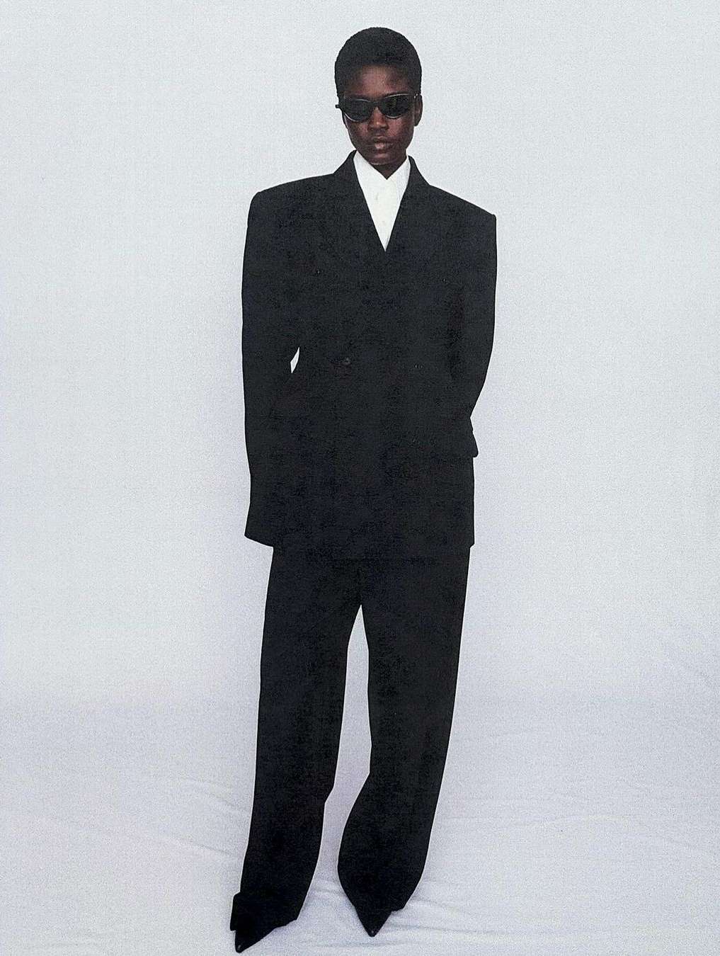 Elegant Black woman in a dark oversized suit and sunglasses stands in a full-length shot against a light backdrop, capturing a fashion-forward aesthetic.