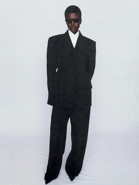Elegant Black woman in a dark oversized suit and sunglasses stands in a full-length shot against a light backdrop, capturing a fashion-forward aesthetic.
