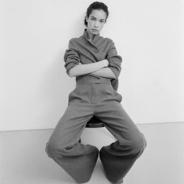 A slender adult female with arms crossed, sits on a stool indoors, wearing a grey jacket and flared trousers in a fashion pose.