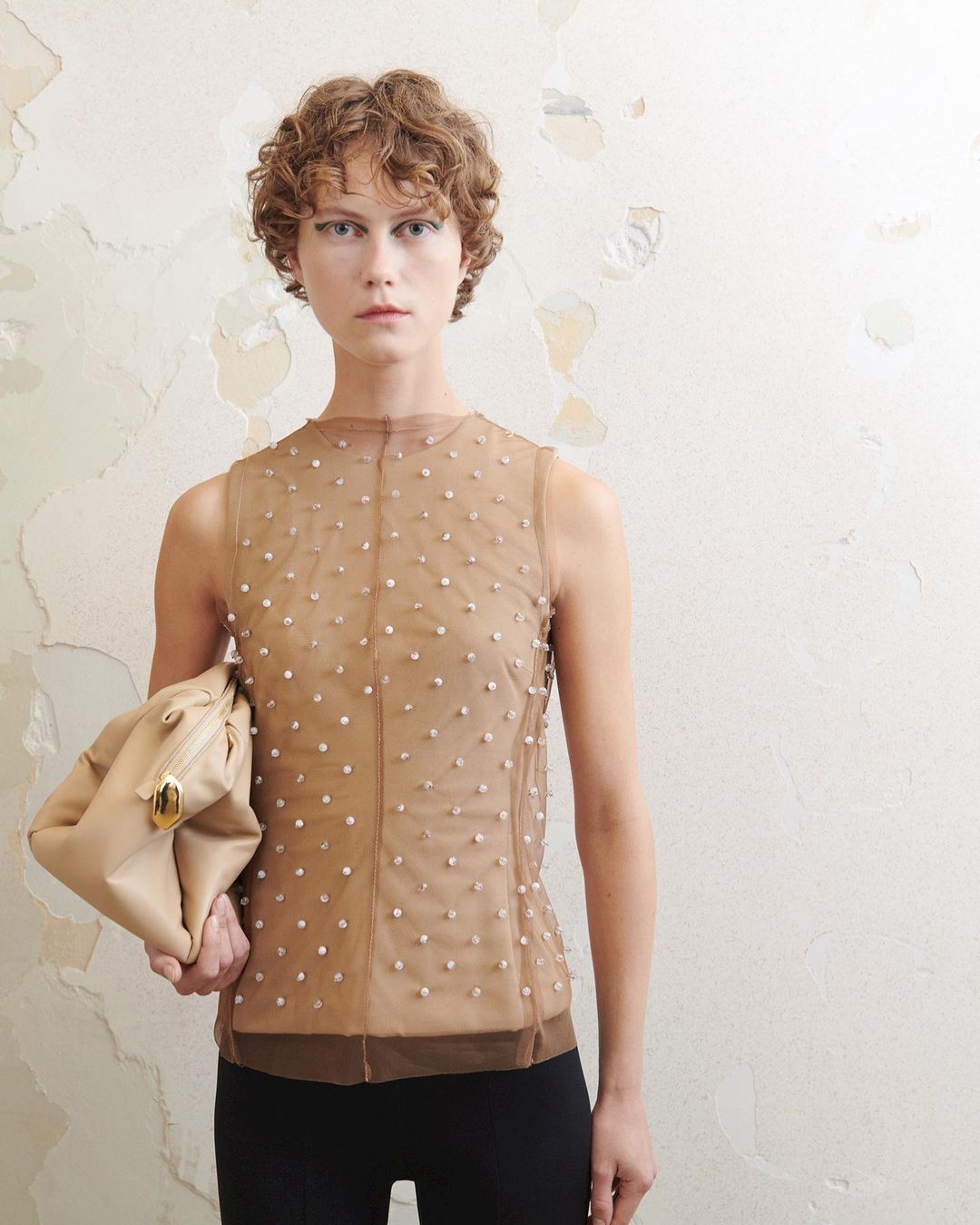Woman in a beige sleeveless top with embellished detail holding a beige clutch against a textured beige wall in a studio.