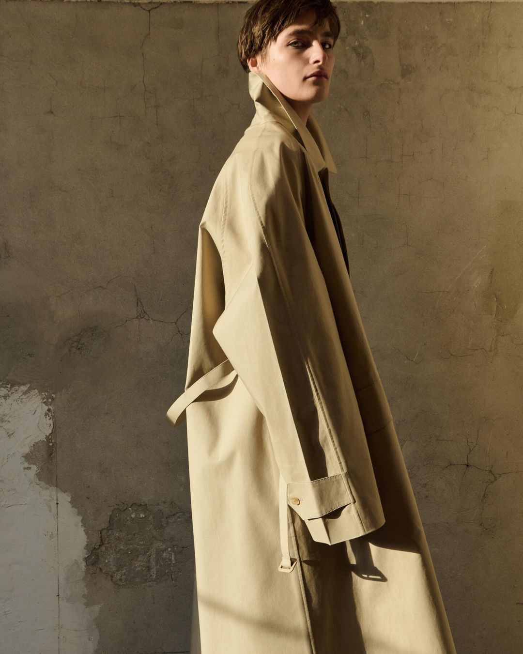 Woman wearing a beige trench coat standing indoors against a textured wall in warm, diffused light. Fashion editorial photography.
