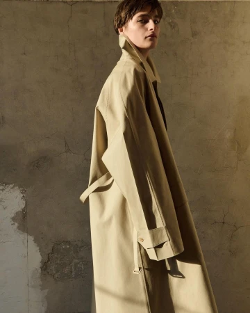 Woman wearing a beige trench coat standing indoors against a textured wall in warm, diffused light. Fashion editorial photography.