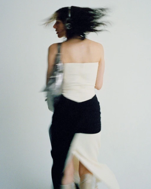 Stylish adult female in motion wearing a strapless top, skirt, boots, and carrying a metallic bag against a white backdrop.