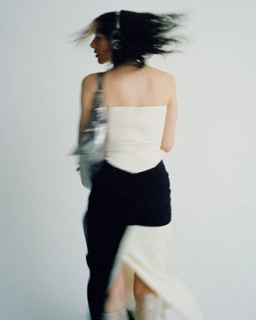Stylish adult female in motion wearing a strapless top, skirt, boots, and carrying a metallic bag against a white backdrop.